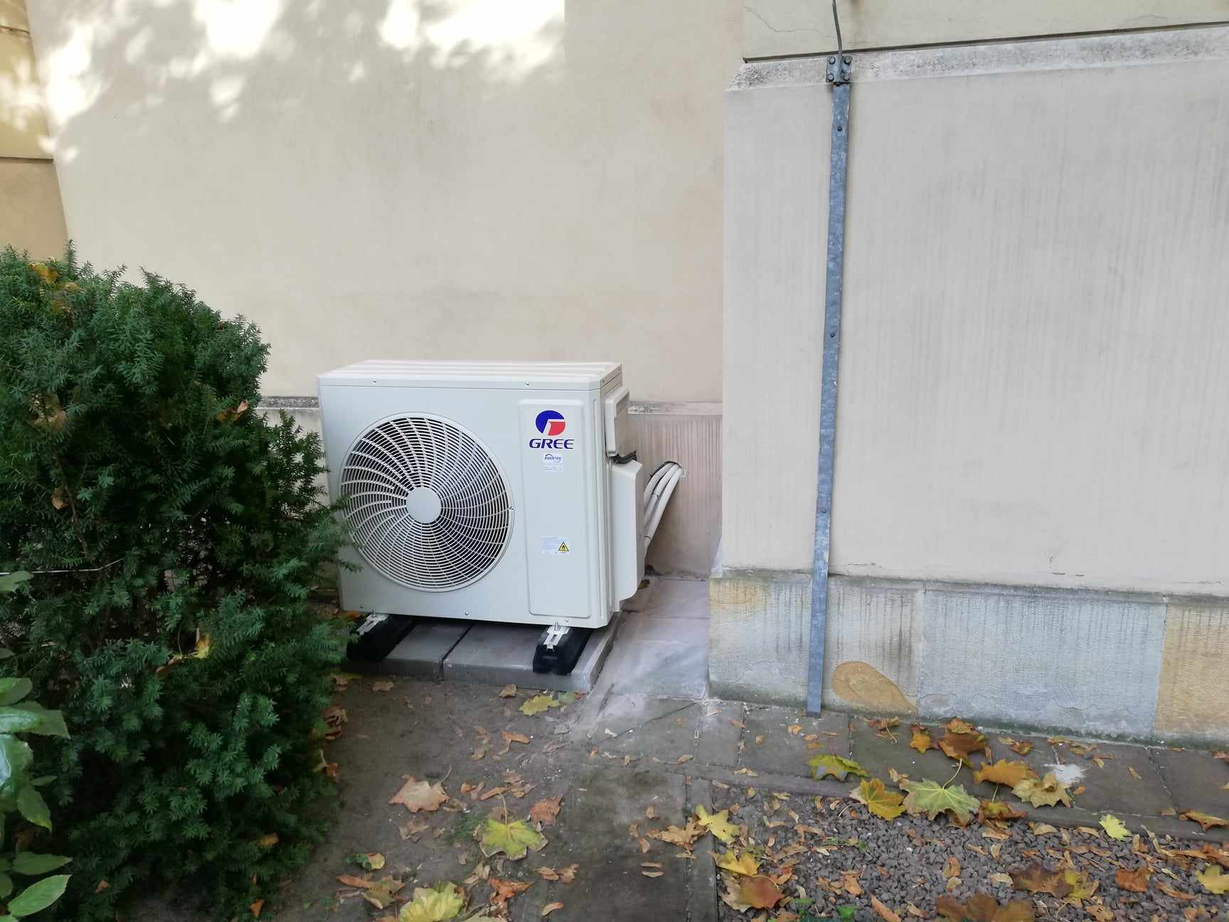 Zewnętrzna jednostka klimatyzacji marki Gree, zamontowana na betonowych płytach przy ścianie budynku, obok krzewu iglastego i opadłych liści.