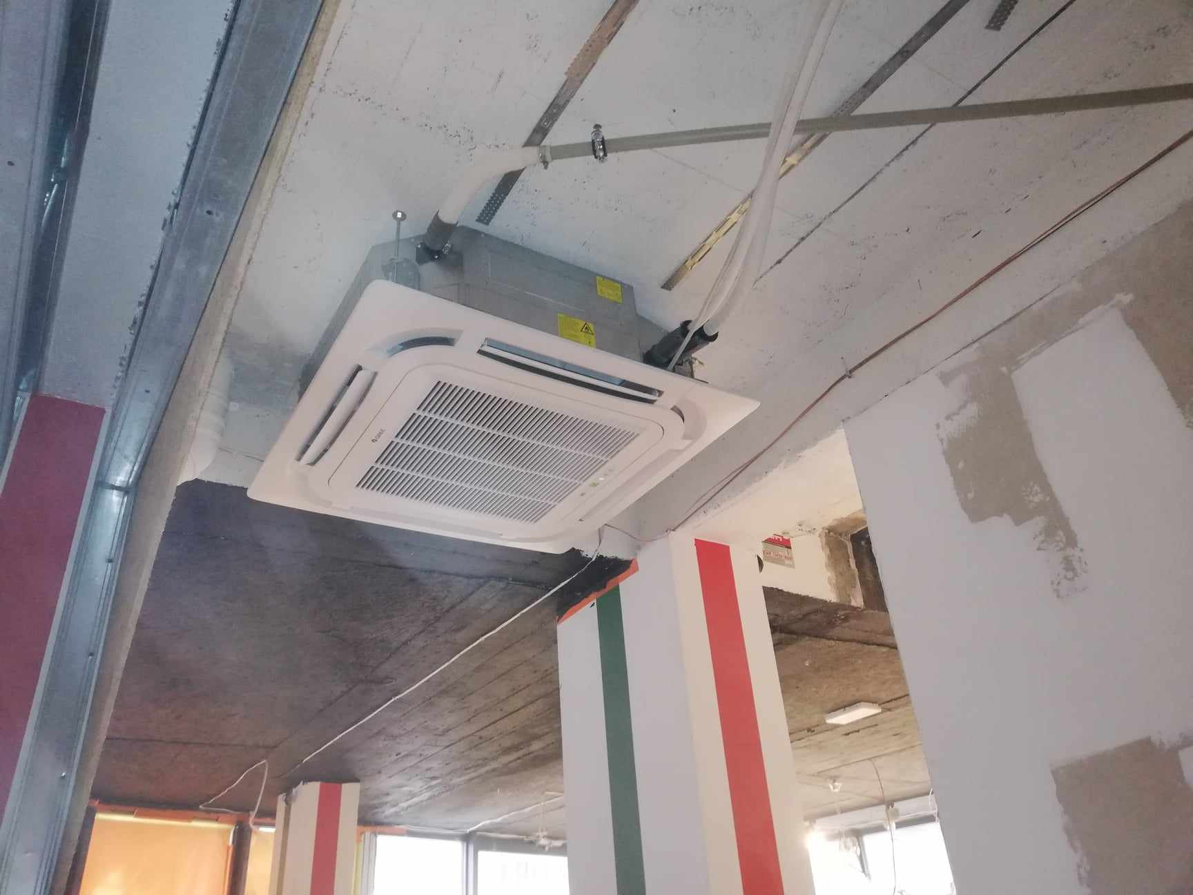 Sufitowa jednostka klimatyzacyjna kasetonowa zamontowana w surowym wnętrzu, widoczne przewody i elementy konstrukcyjne sufitu podwieszanego, kolumna z dekoracyjnymi pasami.