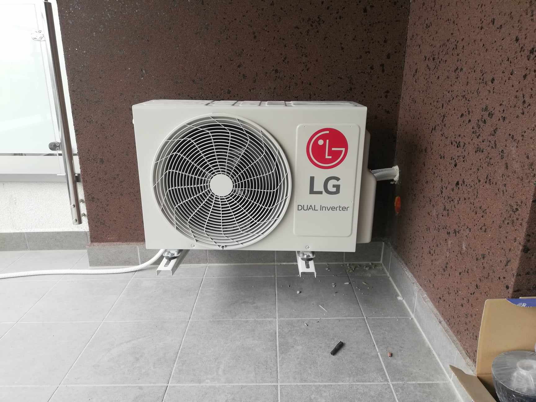 Zewnętrzna jednostka klimatyzatora LG Dual Inverter zamontowana na szarych płytkach balkonu, widoczna biała rurka odprowadzająca skropliny, brązowa ściana w tle.