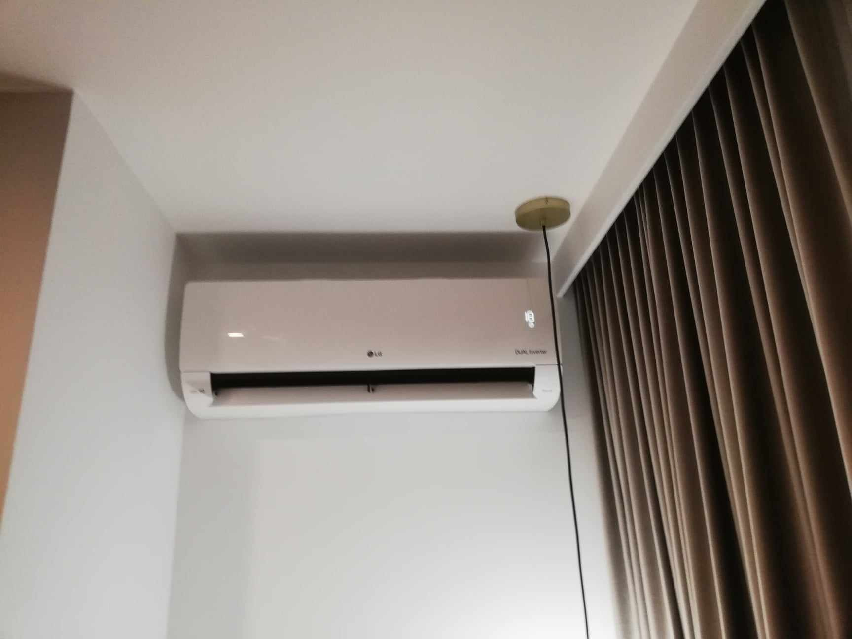 Biały klimatyzator LG Dual Inverter zamontowany na ścianie w pomieszczeniu z beżowymi zasłonami i okrągłą lampą sufitową.