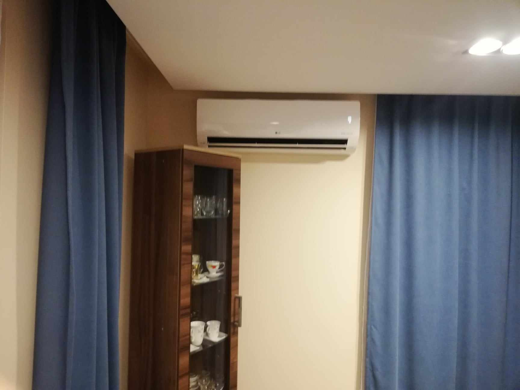 Biała klimatyzacja ścienna LG zamontowana nad drewnianą witryną z szklanymi drzwiami, obok zasłony w kolorze denim, w pokoju o beżowych ścianach.