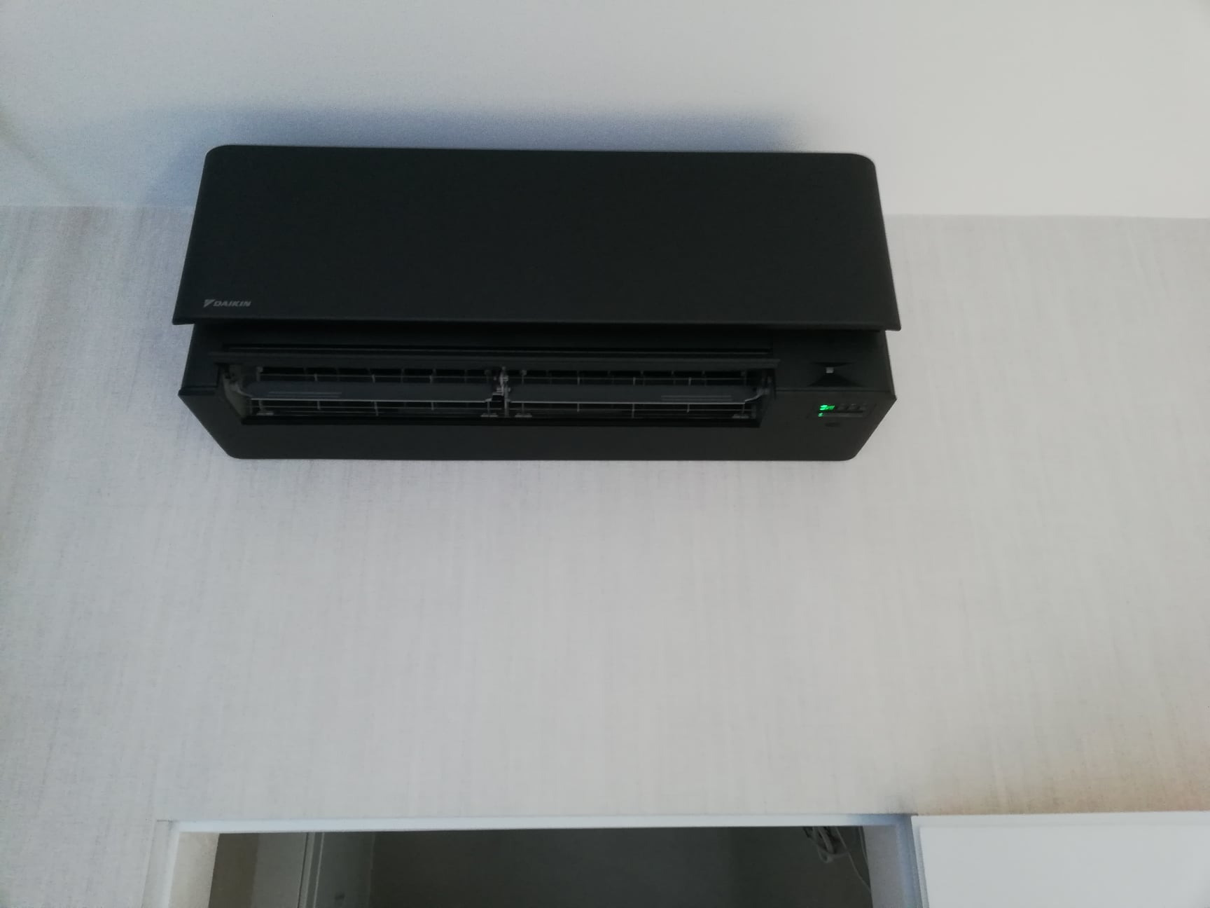 Czarna jednostka wewnętrzna klimatyzacji Daikin zamontowana na jasnej, wzorzystej ścianie. Widoczna zielona dioda sygnalizacyjna.