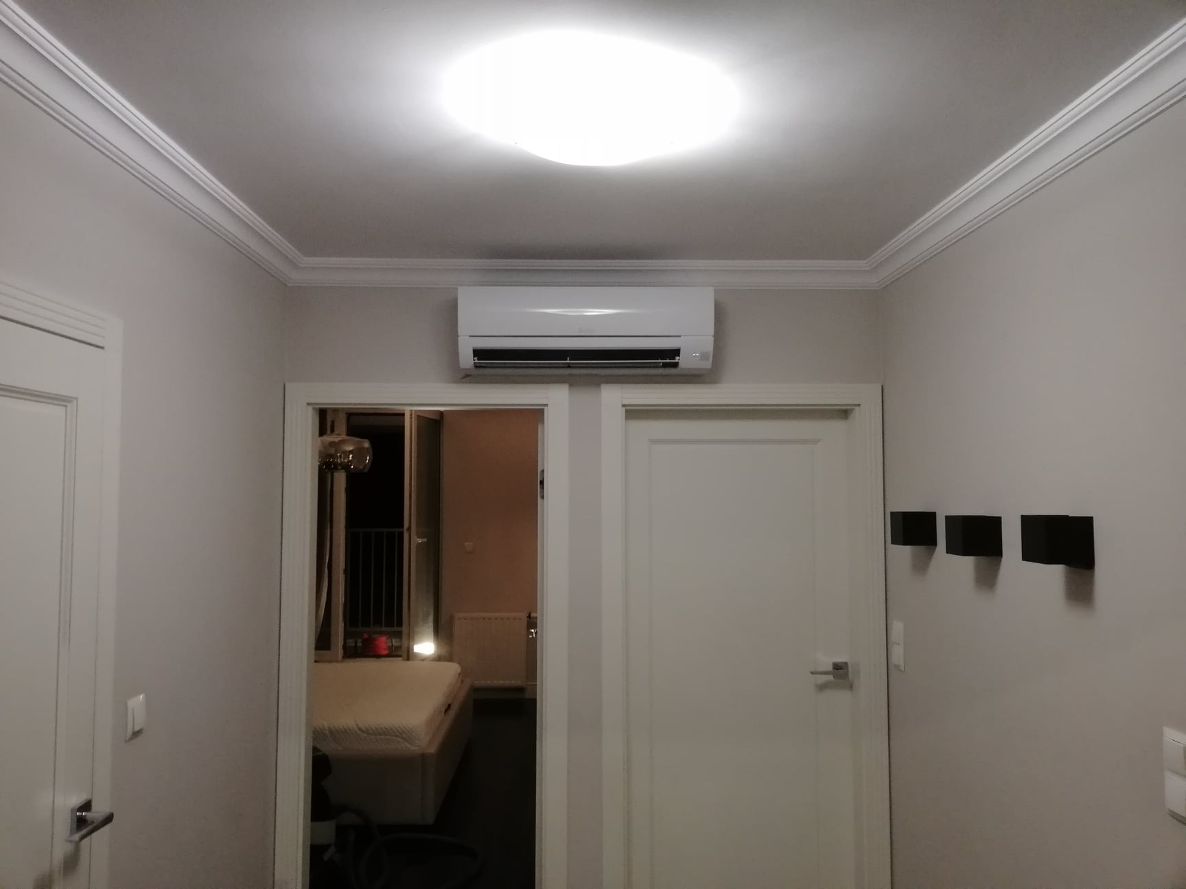 Biały klimatyzator ścienny zamontowany nad drzwiami w jasnym pokoju z dekoracyjną listwą przysufitową.