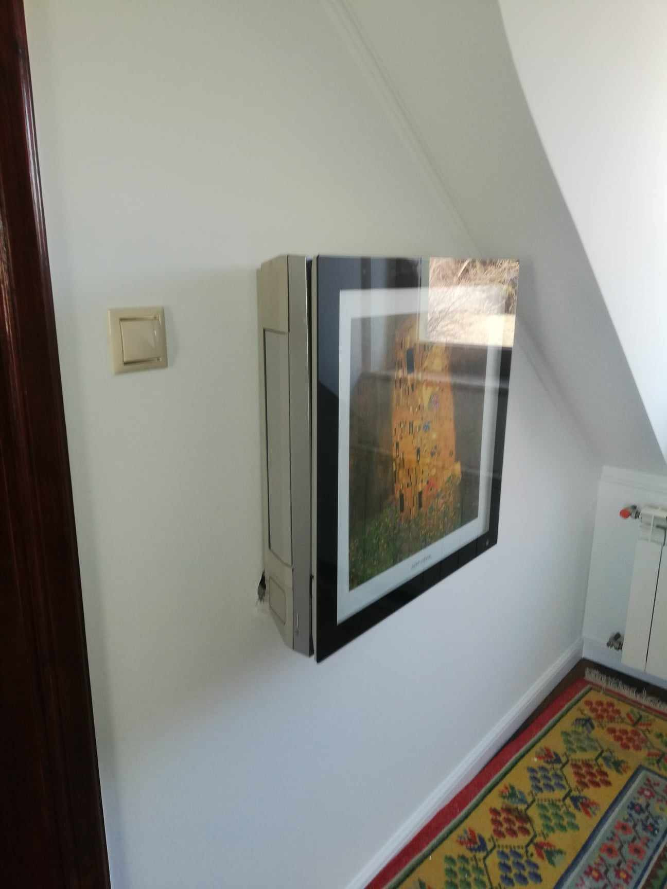 Klimatyzator ścienny zamontowany na ścianie pod skosem, obok włącznika światła, z obrazem Gustava Klimta w ramie na froncie, na tle kolorowego dywanu.