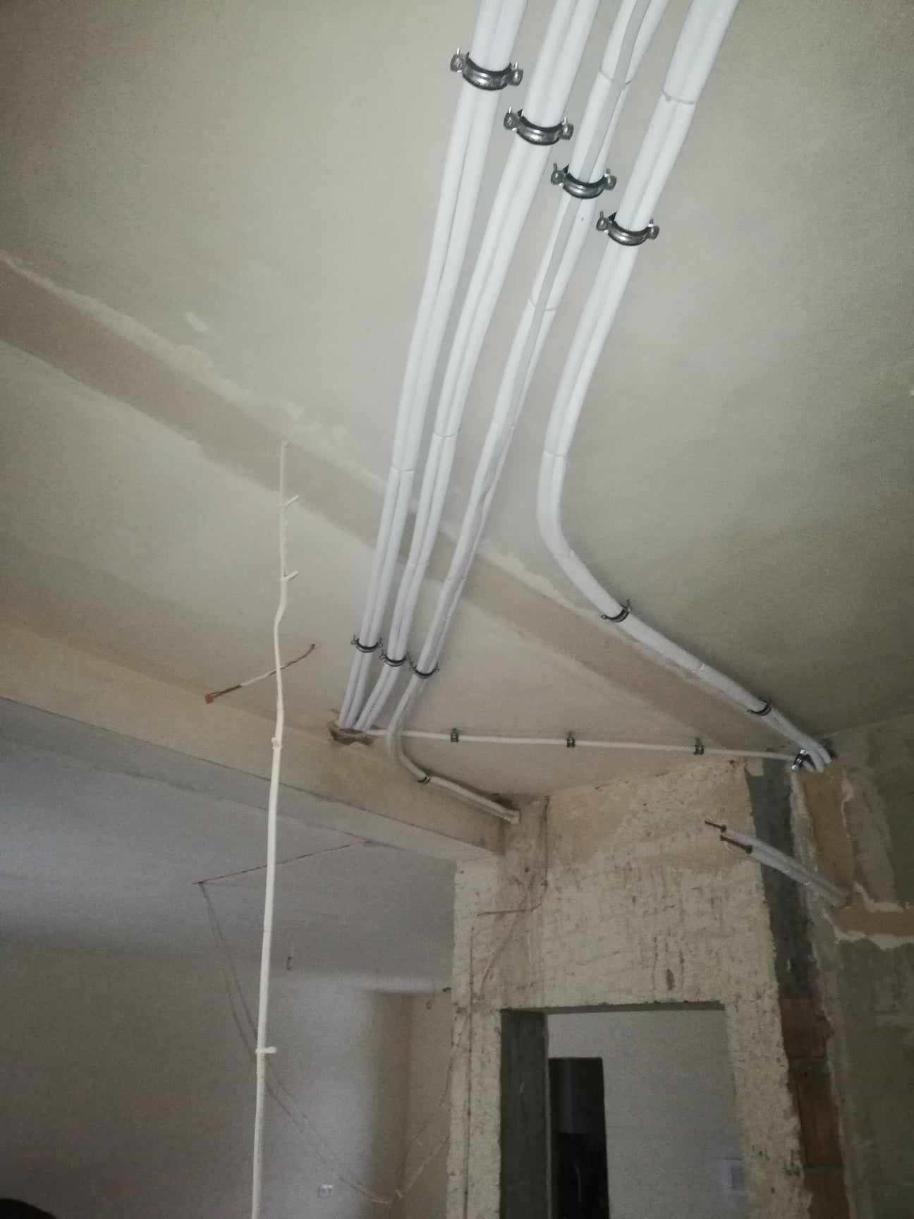 Białe rury wentylacyjne przymocowane metalowymi uchwytami do sufitu w surowym stanie deweloperskim, widoczne fragmenty ścian z odsłoniętą cegłą i niedokończone tynki.