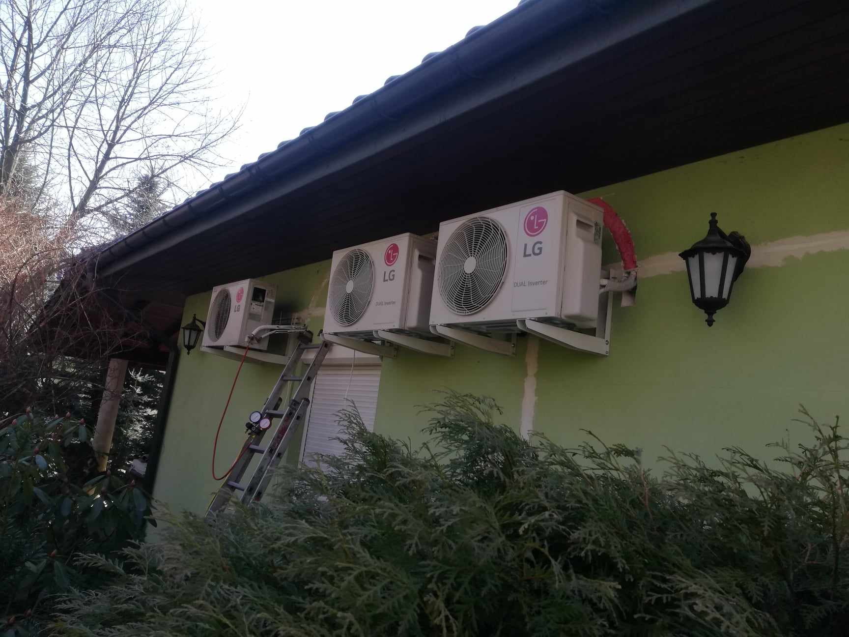 Trzy jednostki zewnętrzne klimatyzacji LG zamontowane na zielonej ścianie domu, widoczna drabina z manometrami, dekoracyjna lampa zewnętrzna.