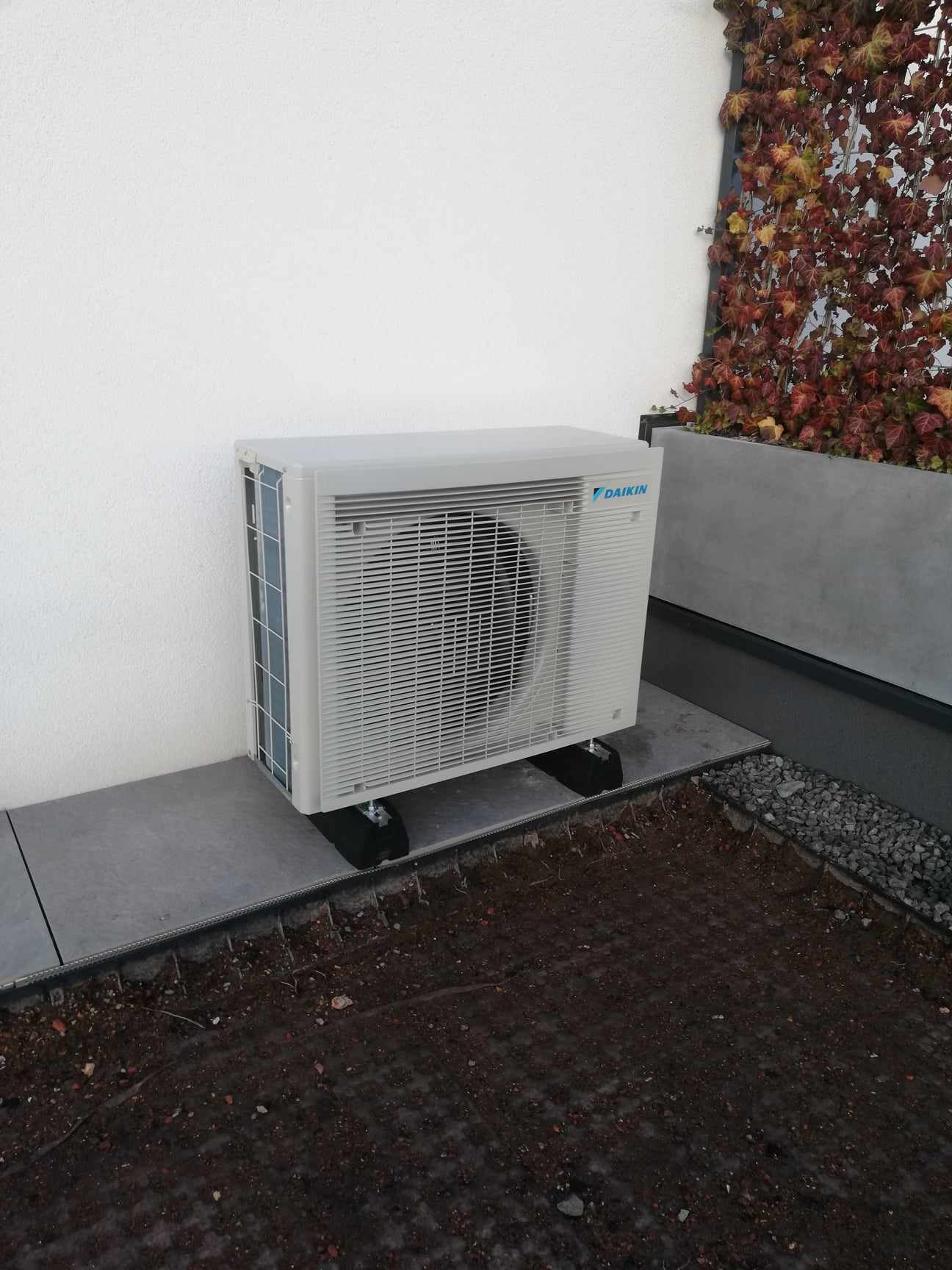 Zewnętrzna jednostka klimatyzacji Daikin zamontowana na szarych płytach w Przegini, z bluszczem w tle, widoczne czarne nóżki podtrzymujące urządzenie.