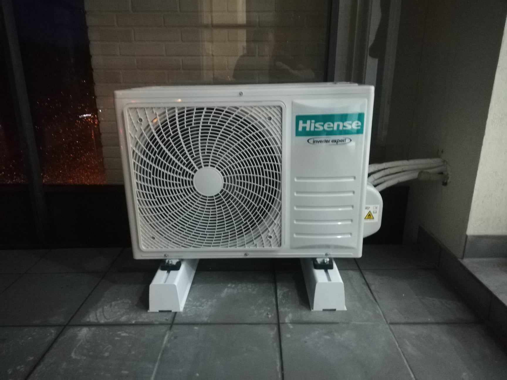 Biała jednostka zewnętrzna klimatyzacji Hisense Inverter Expert, umieszczona na wspornikach na szarym balkonie, z widocznymi przewodami wychodzącymi ze ściany.