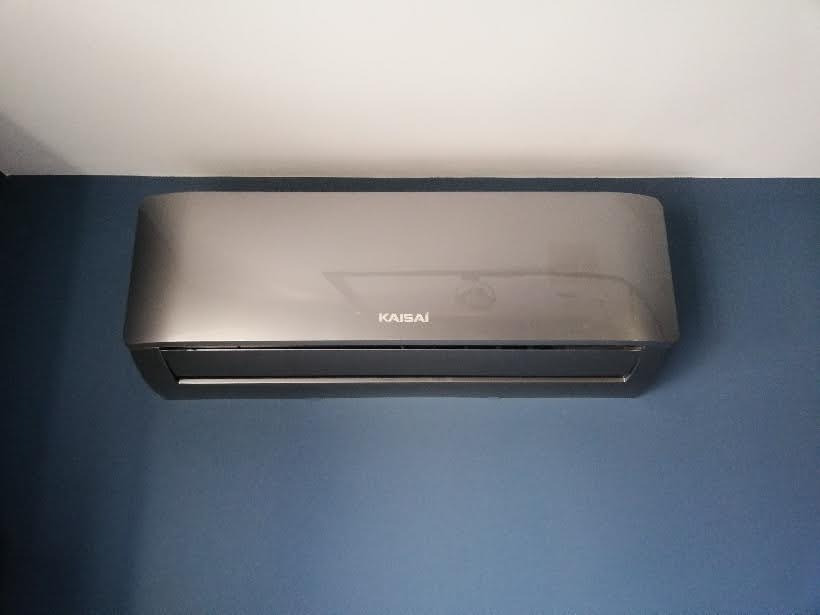 Szary klimatyzator marki Kaisai zamontowany na niebieskiej ścianie, widok z dołu.