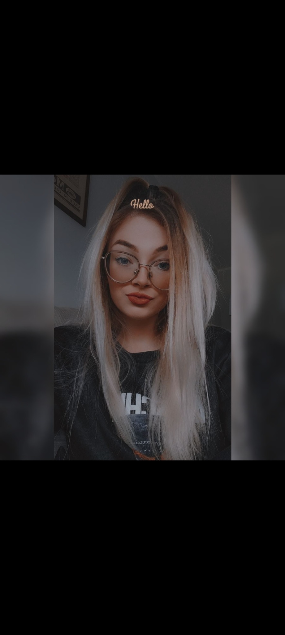 Portret kobiety z blond włosami w okularach, z napisem 'Hello' na włosach, w ciemnym pomieszczeniu.