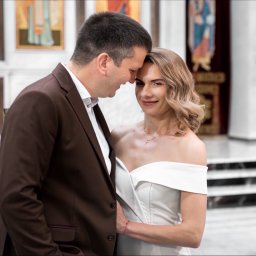 Ernest Machalski Fotografia - Para młoda w kościele: on w brązowym garniturze, ona w białej sukni, delikatny uśmiech i bliskość. W tle ikony i marmurowe schody.