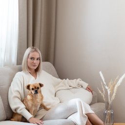 Ernest Machalski Fotografia - Kobieta z krótkimi blond włosami w kremowym swetrze siedzi na beżowej kanapie z brązowym psem; obok stolik z dekoracyjnym bukietem w wazonie, stonowane wnętrze.