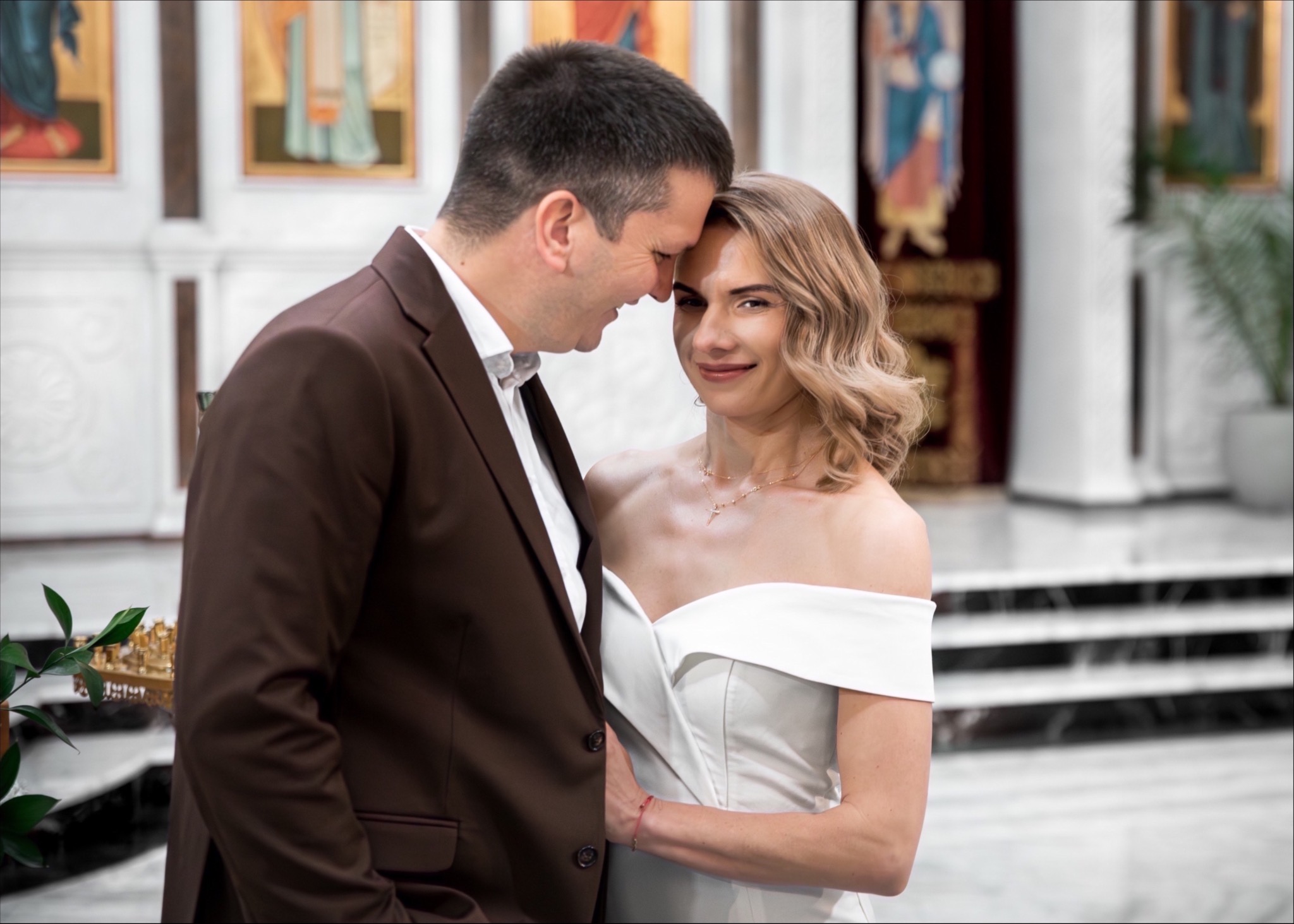 Para młoda w kościele: on w brązowym garniturze, ona w białej sukni, delikatny uśmiech i bliskość. W tle ikony i marmurowe schody.