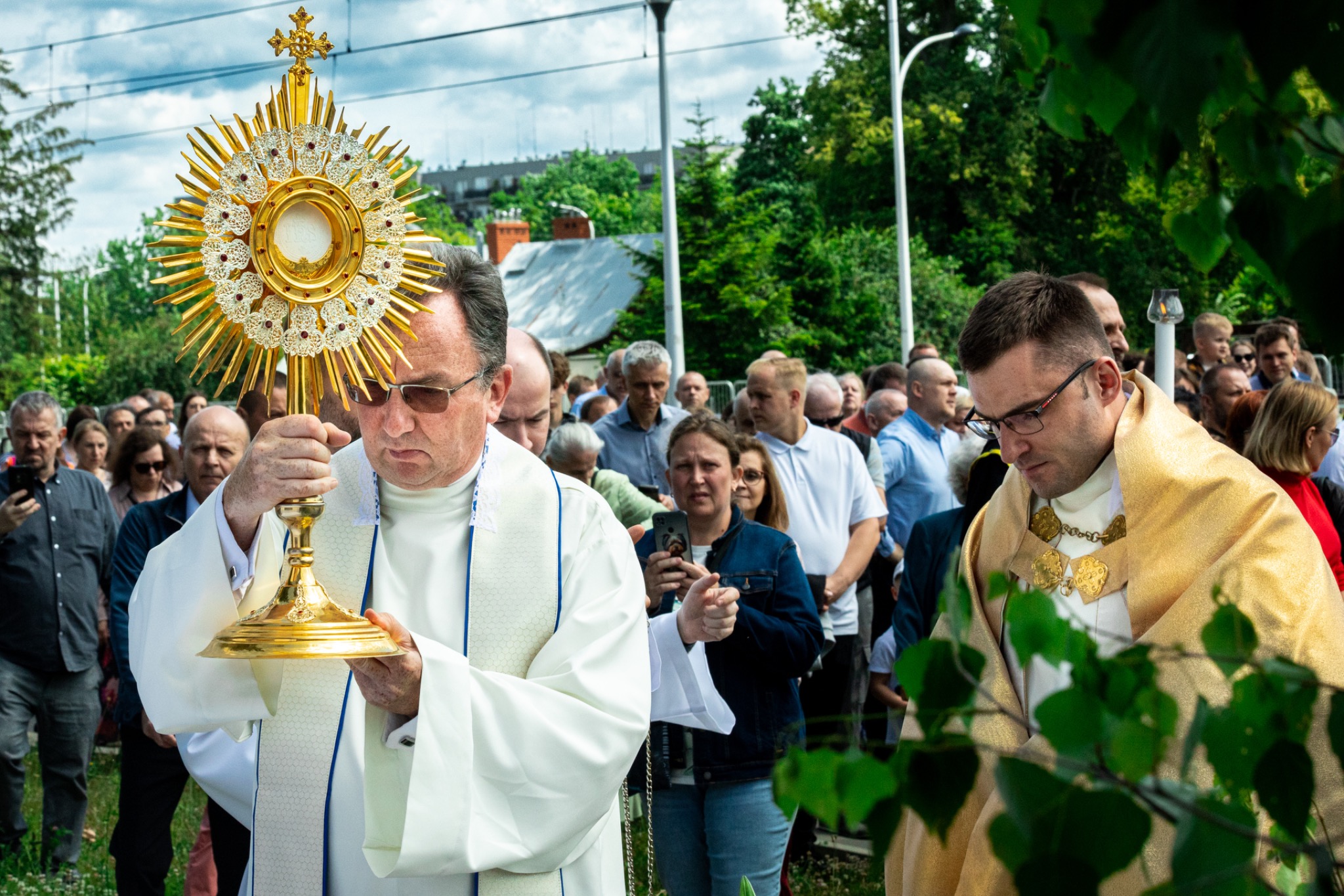 Procesja Bożego Ciała w Warszawie: duchowni niosą monstrancję, tłum wiernych w tle. Uroczystość religijna w plenerze, słoneczny dzień, zielone otoczenie.