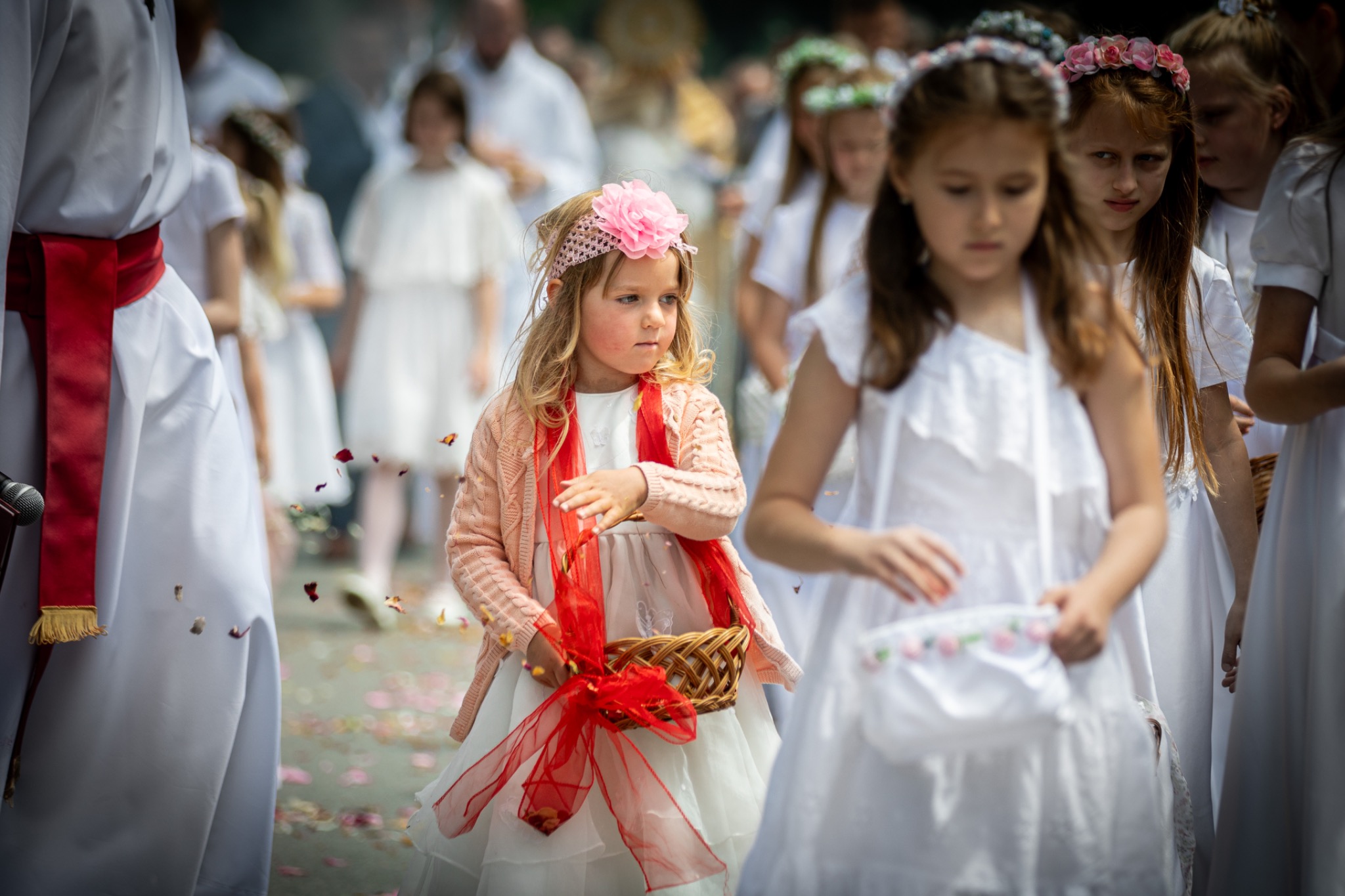 Dziewczynka w białej sukience z różową opaską na głowie i koszyczkiem z płatkami kwiatów podczas procesji komunijnej. W tle inne dzieci w białych strojach.