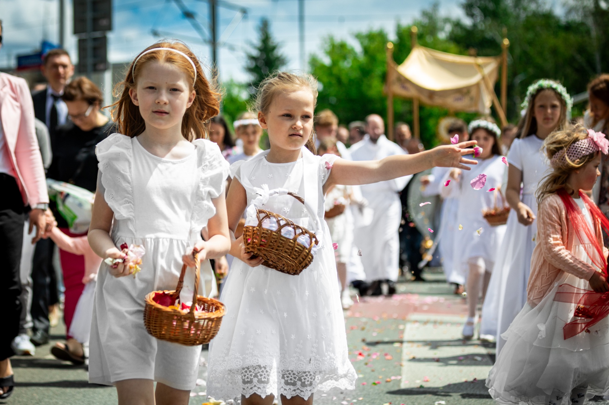 Procesja Bożego Ciała: dziewczynki w białych sukienkach sypią płatki róż, niosąc wiklinowe koszyczki. W tle baldachim i uczestnicy procesji.