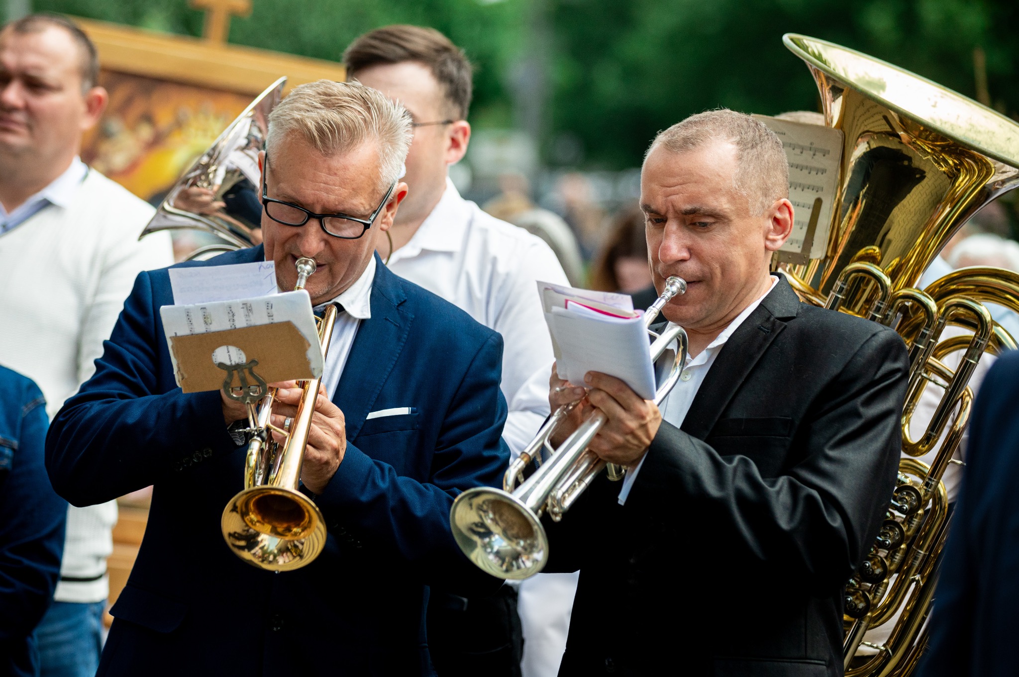 Dwóch mężczyzn w garniturach gra na trąbkach, obok nich tuba. Zespół muzyczny podczas uroczystości. Widoczne nuty na statywach.