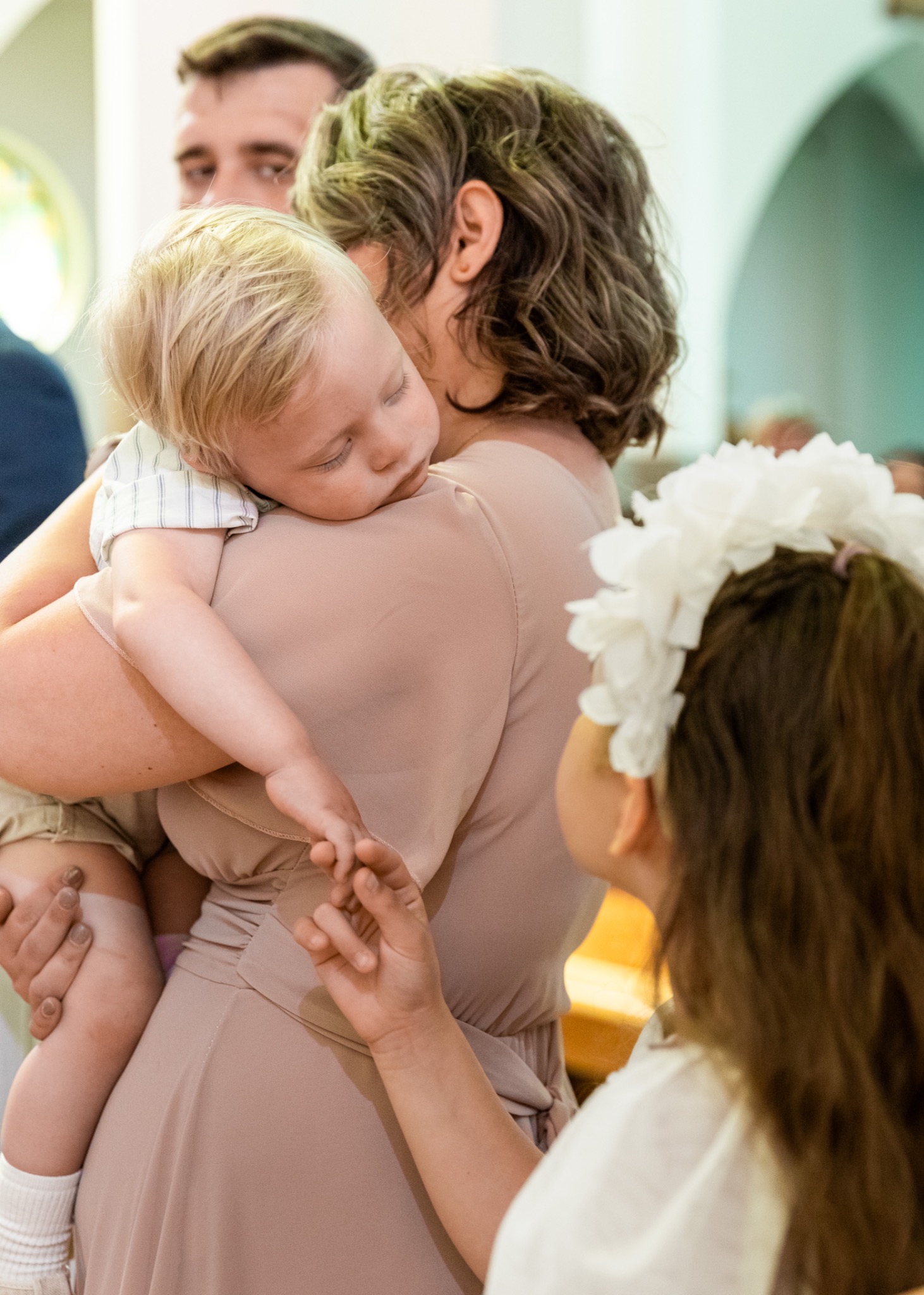 Matka trzyma śpiącego blond chłopca na rękach, obok dziewczynka z wiankiem. Ujęcie z uroczystości religijnej, w tle widoczne okno z witrażem.