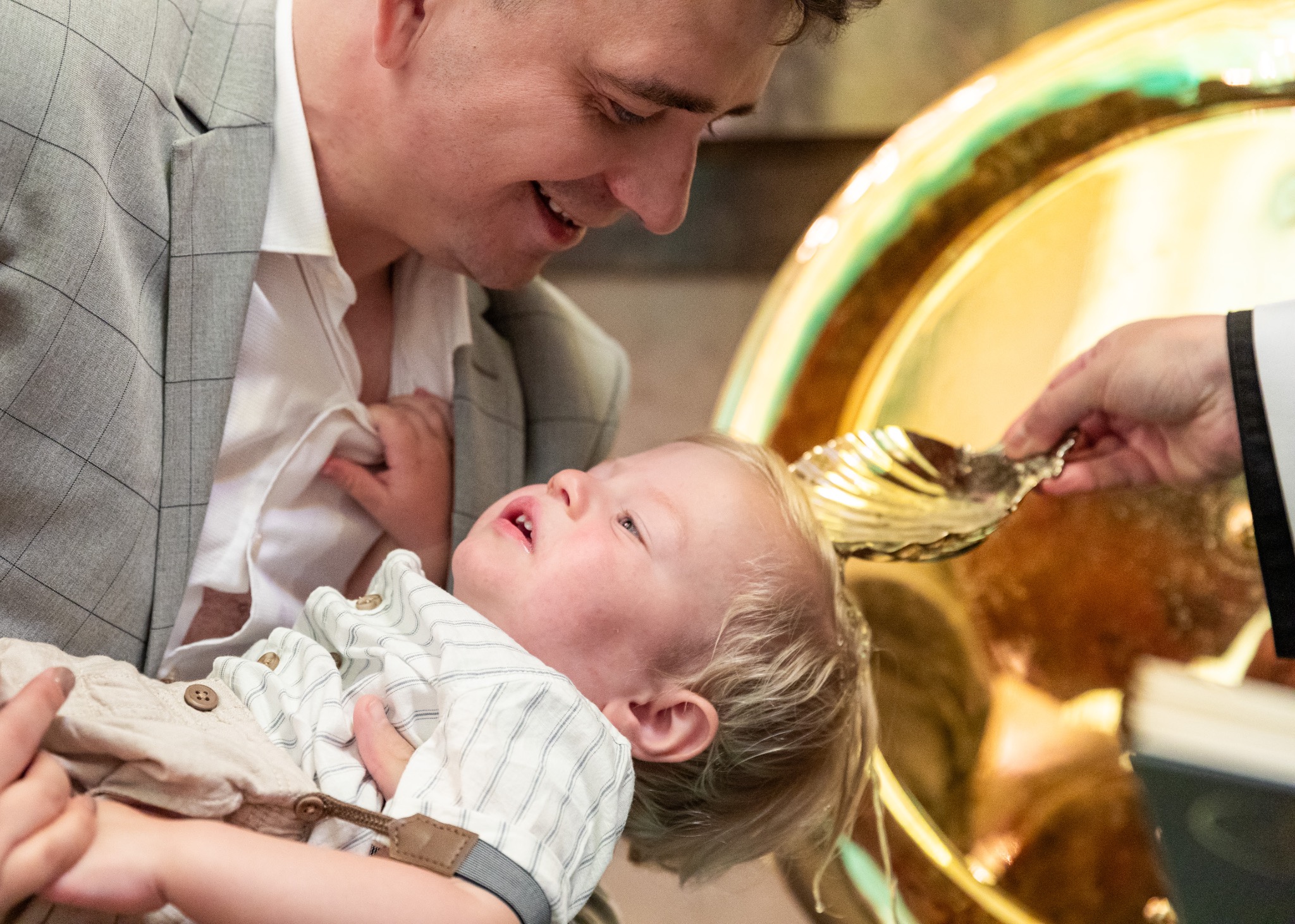 Uśmiechnięty ojciec trzyma na rękach małego chłopca podczas chrztu, w tle złota misa i dłoń księdza z muszlą. Jasne, naturalne światło.