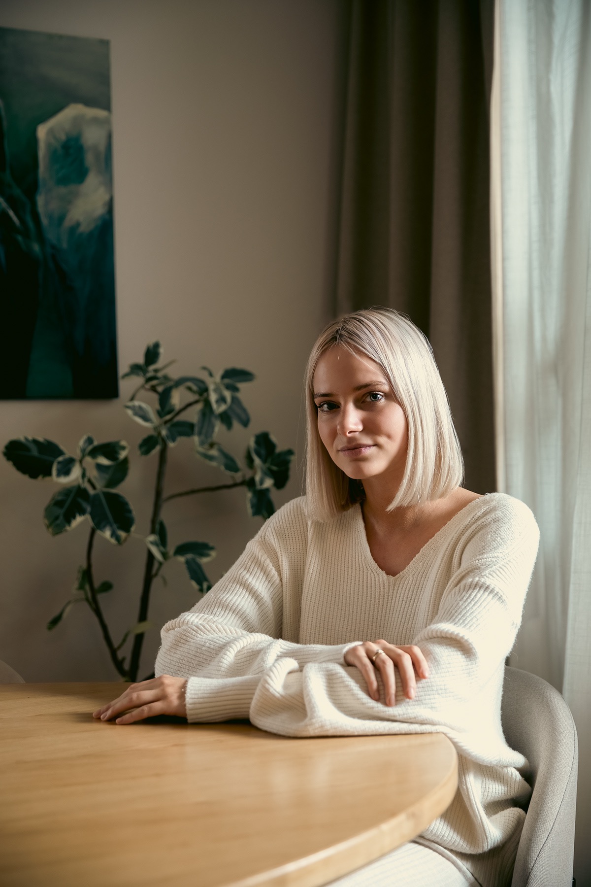 Portret kobiety o platynowych włosach w kremowym swetrze, siedzącej przy okrągłym, drewnianym stole w minimalistycznym wnętrzu z obrazem i rośliną w tle.