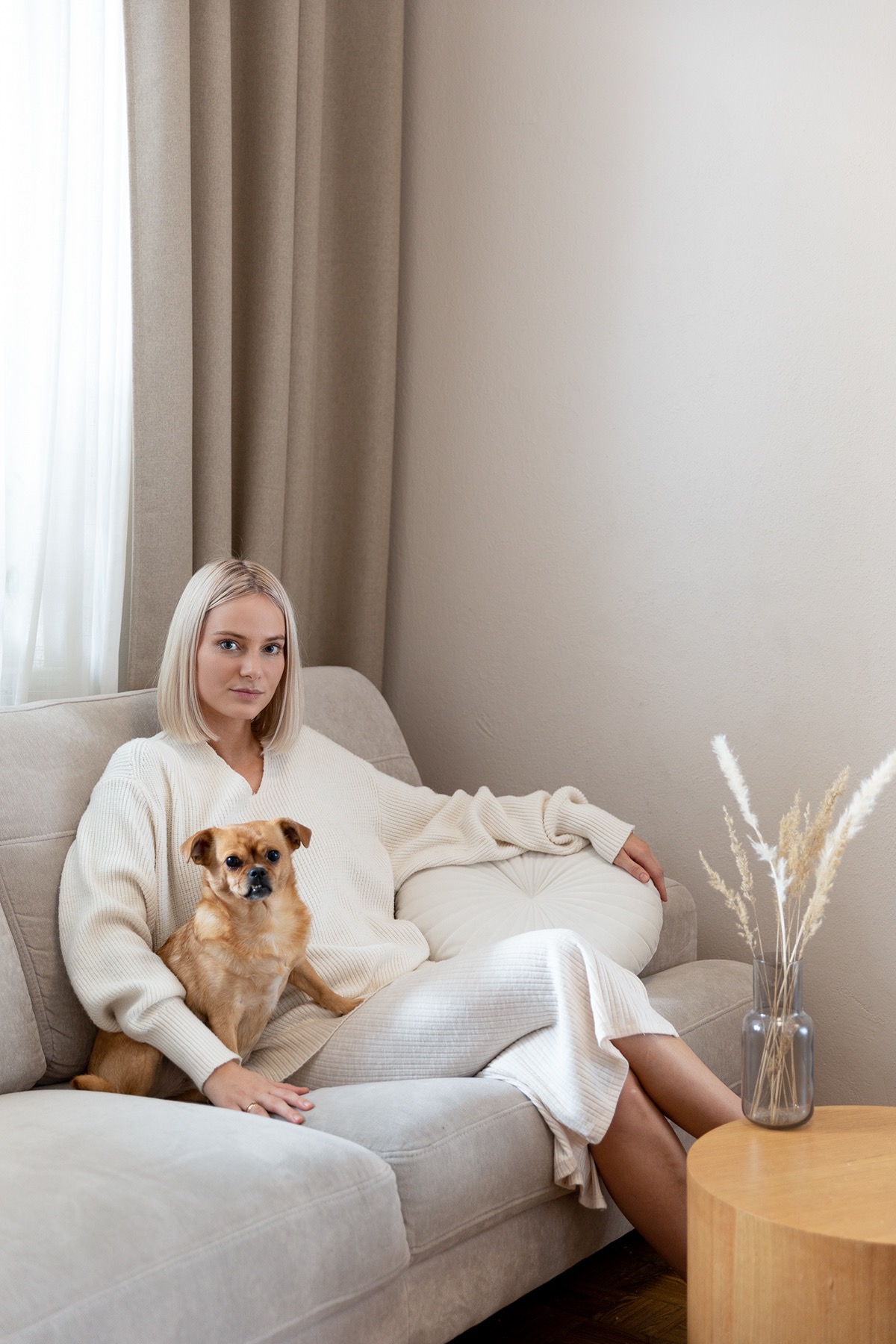 Kobieta z krótkimi blond włosami w kremowym swetrze siedzi na beżowej kanapie z brązowym psem; obok stolik z dekoracyjnym bukietem w wazonie, stonowane wnętrze.