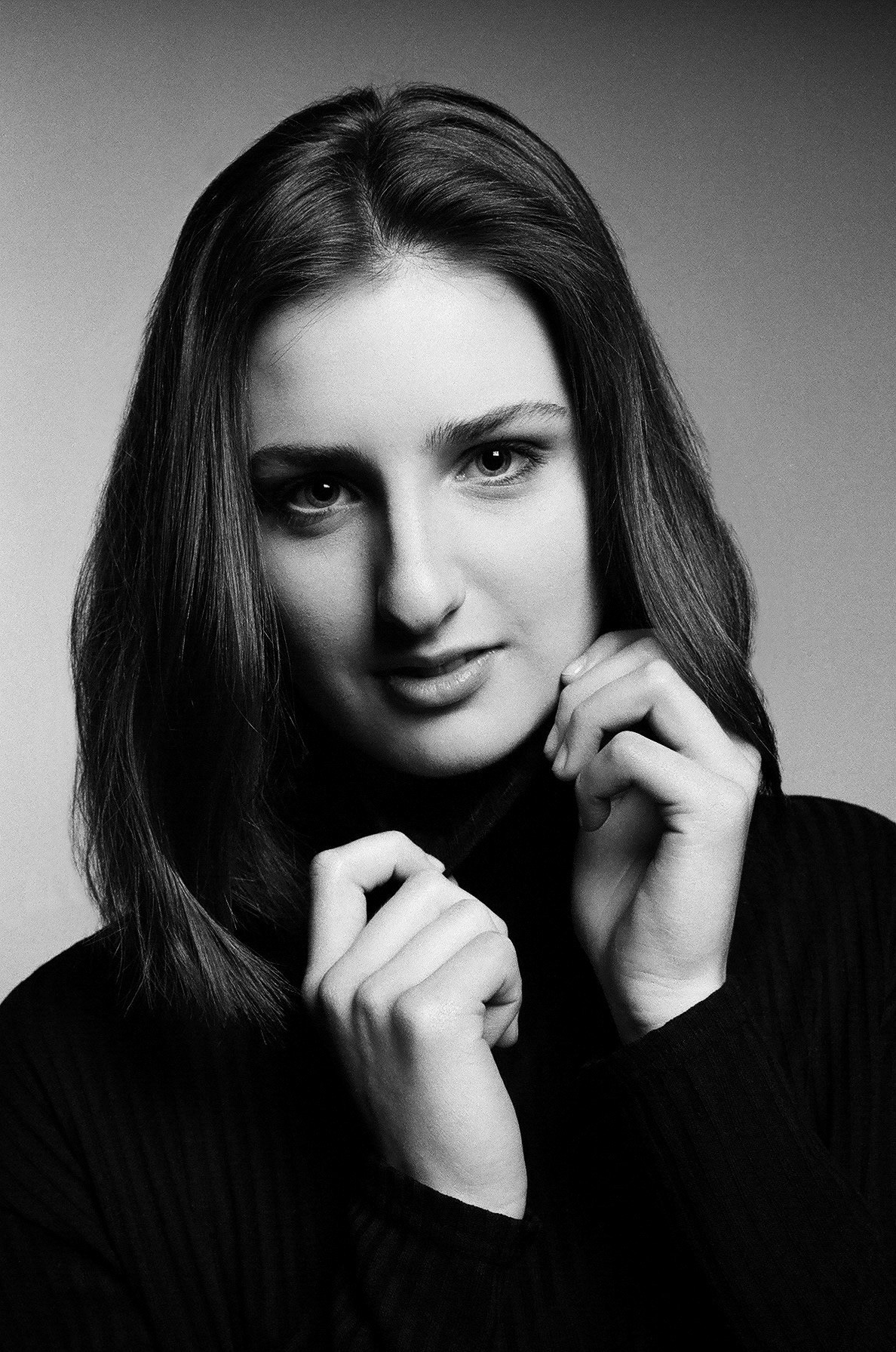 Portret młodej kobiety w czarnym golfie, czarno-białe zdjęcie z subtelnym uśmiechem i dłońmi przy szyi.