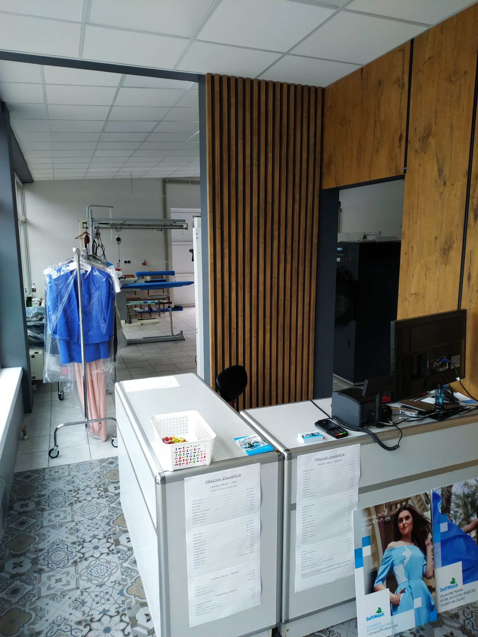 Wnętrze pralni chemicznej: wieszak z niebieskimi ubraniami w foliowych pokrowcach, stół do prasowania w tle, lada recepcyjna z cennikiem usług i reklamą, posadzka z wzorem geometrycznym.