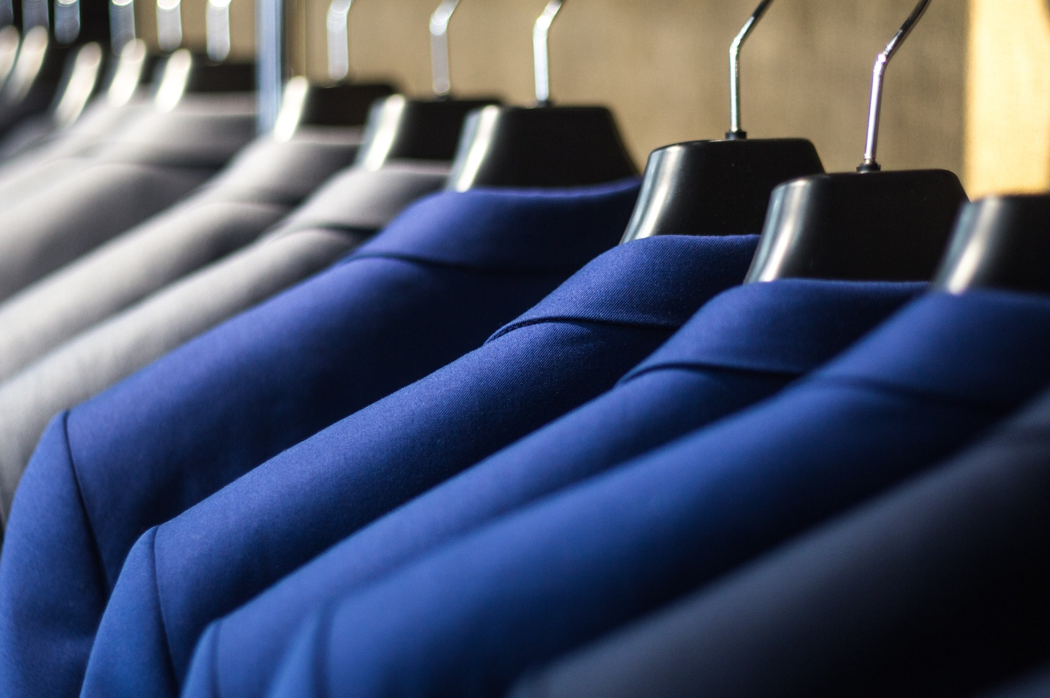 Rząd marynarek w odcieniach szarości i błękitu wiszących na czarnych wieszakach, widoczne z bliska, z ostrością na błękitne tkaniny.