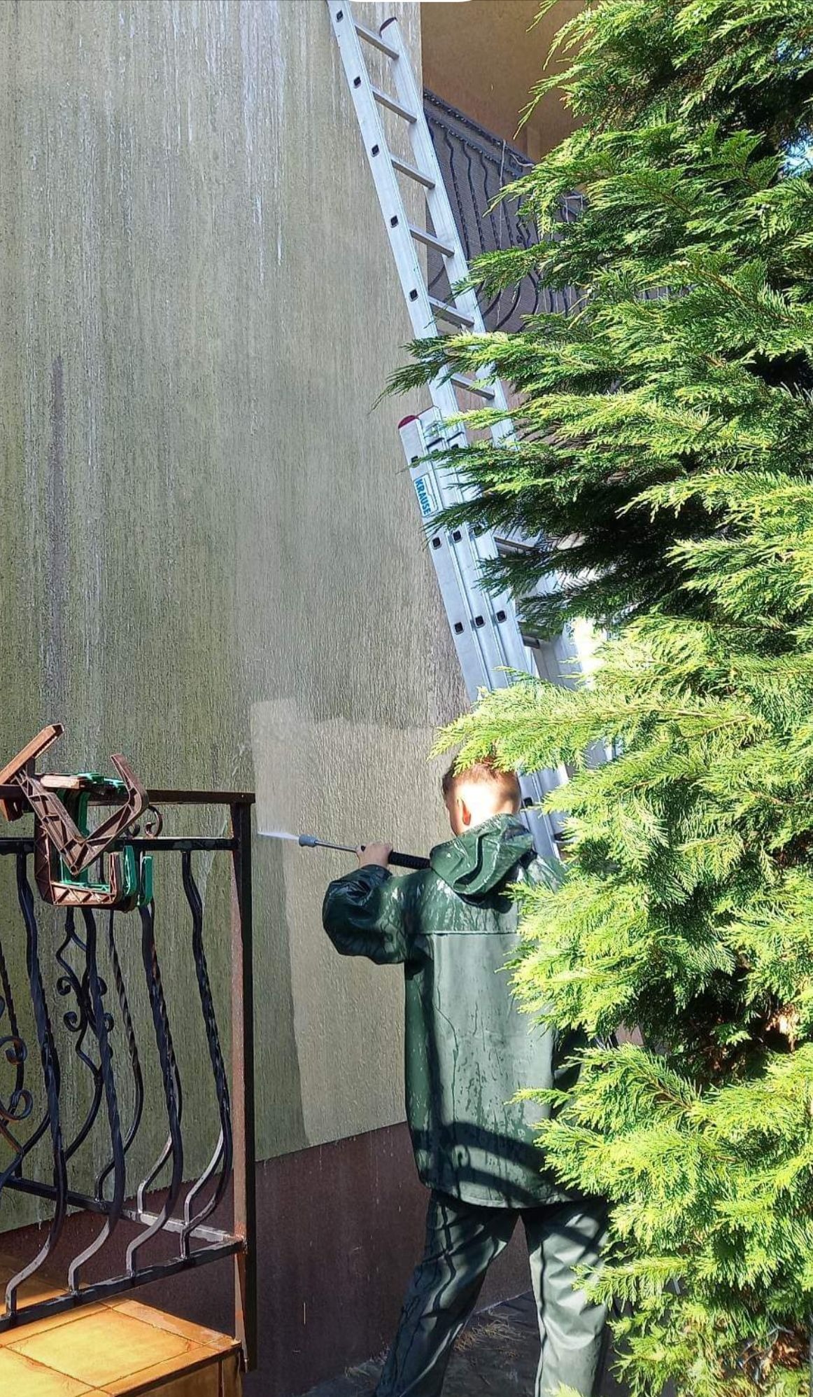 Pracownik w zielonym kombinezonie spryskuje ścianę budynku myjką ciśnieniową, widoczny fragment wyczyszczonej powierzchni, drabina oparta o ścianę, obok rośnie iglak.