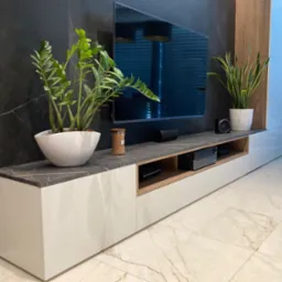Nowoczesna szafka RTV z białymi frontami i blatem z ciemnego marmuru, uzupełniona roślinami w białych donicach i czarnym telewizorem na tle ściany z czarnego marmuru.