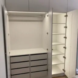 Wnętrze białej szafy wnękowej z otwartymi drzwiami, widoczne półki, szuflady i oświetlenie LED. Parkiet w jodełkę na podłodze. Minimalistyczny design.