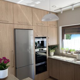 AURA Project Mariusz Kręgiel - Jasna kuchnia z drewnianymi frontami, zabudowana od podłogi do sufitu. Widoczna lodówka, piekarnik, okno z roletami i kwiaty w doniczkach na blacie.