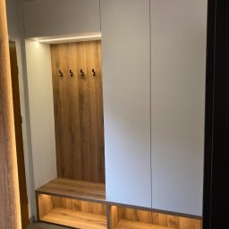 AURA Project Mariusz Kręgiel - Biała szafa wnękowa z drewnianym panelem z haczykami i podświetlaną półką na buty, na tle szarej podłogi, widziana z perspektywy wejścia.