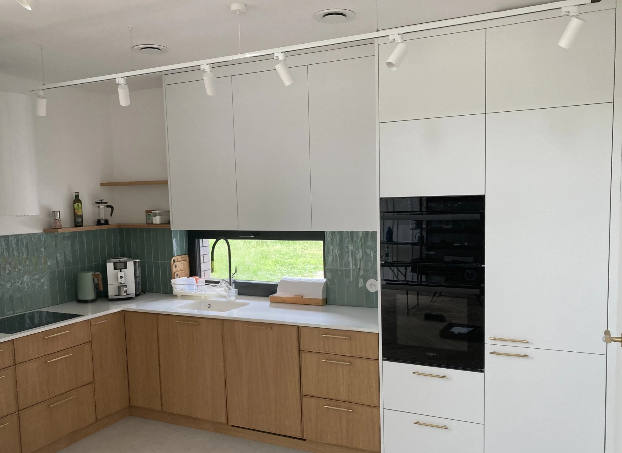 Nowoczesna kuchnia z drewnianymi frontami dolnych szafek i białymi górnymi, oświetlona szynoprzewodem. Widok na zieleń za oknem.