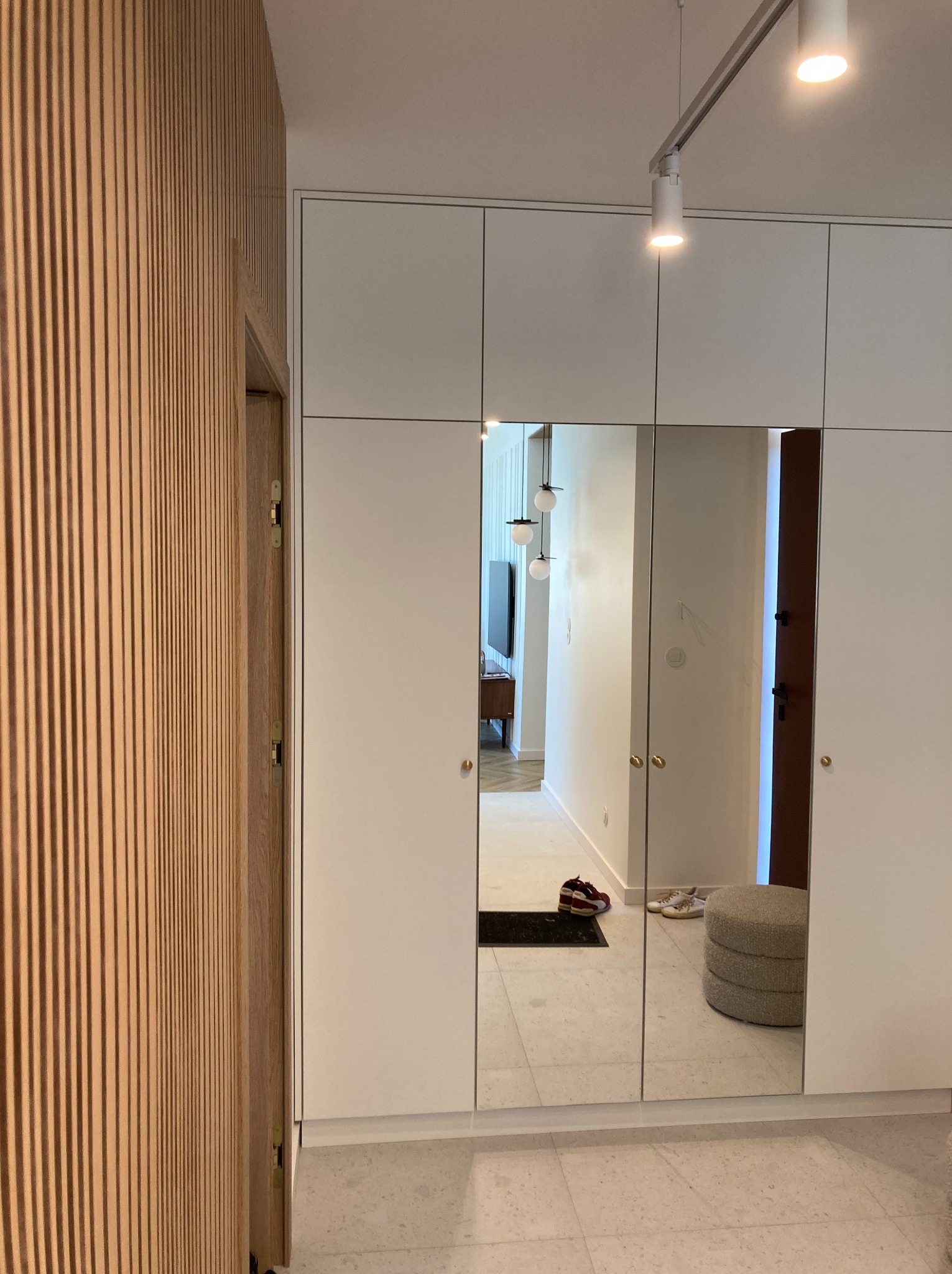 Biała szafa wnękowa z lustrzanymi drzwiami odbija wnętrze mieszkania, obok drewniana ściana z lamelami i lampa sufitowa w nowoczesnym stylu.