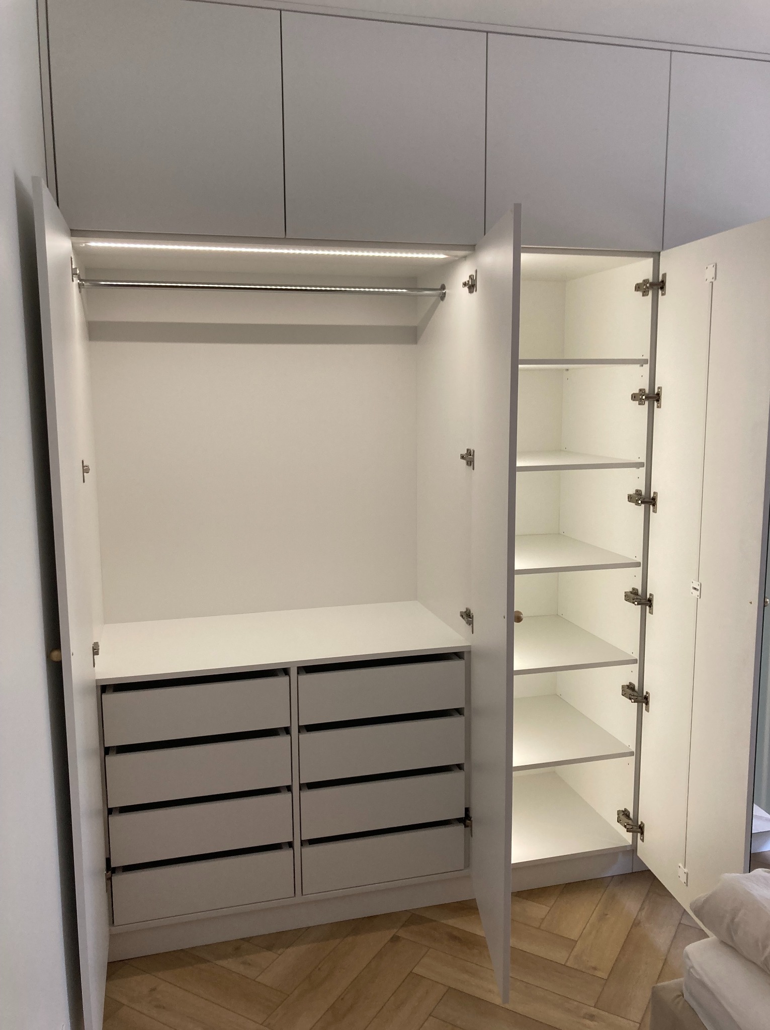 Wnętrze białej szafy wnękowej z otwartymi drzwiami, widoczne półki, szuflady i oświetlenie LED. Parkiet w jodełkę na podłodze. Minimalistyczny design.