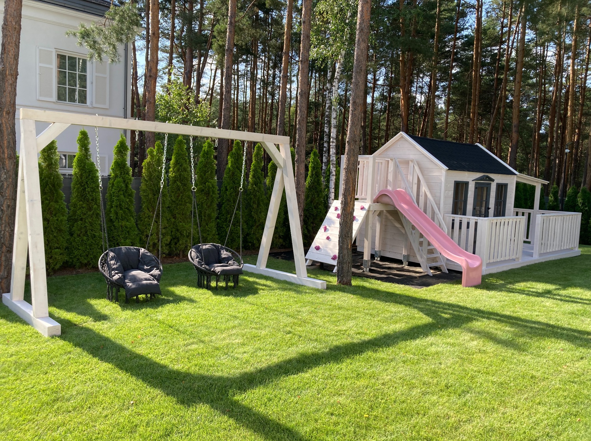 Drewniany plac zabaw z huśtawką-ławka, domkiem ze zjeżdżalnią i ścianką wspinaczkową na zielonym trawniku w ogrodzie. Biała konstrukcja, ciemne poduszki na huśtawce.