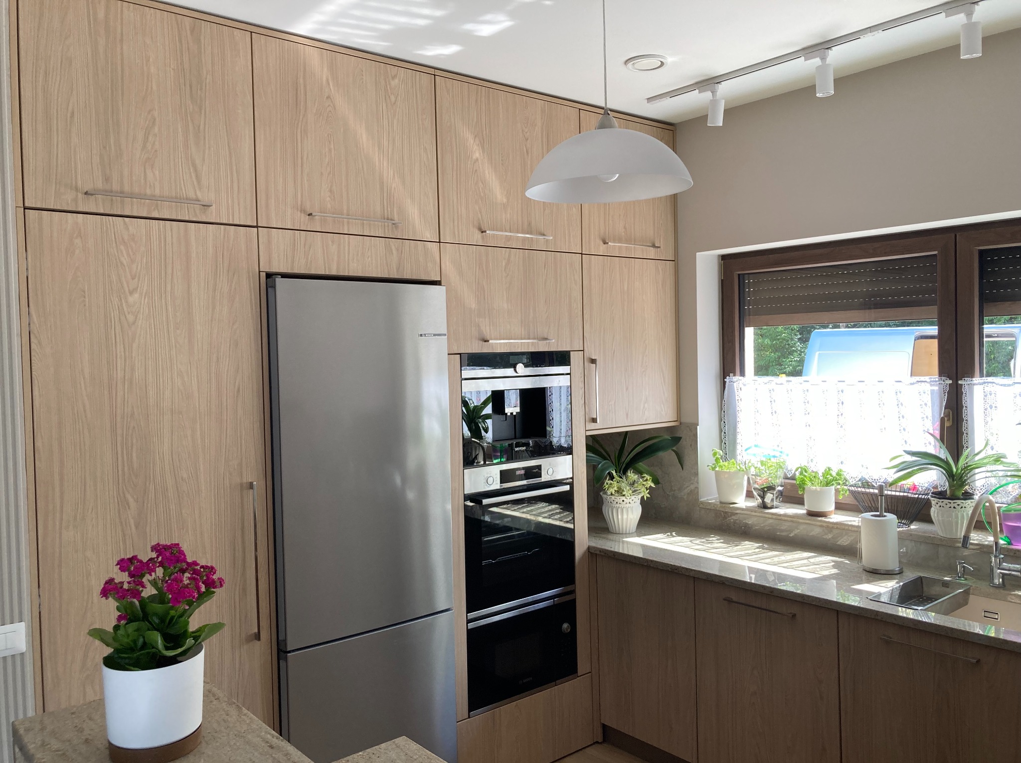 Jasna kuchnia z drewnianymi frontami, zabudowana od podłogi do sufitu. Widoczna lodówka, piekarnik, okno z roletami i kwiaty w doniczkach na blacie.