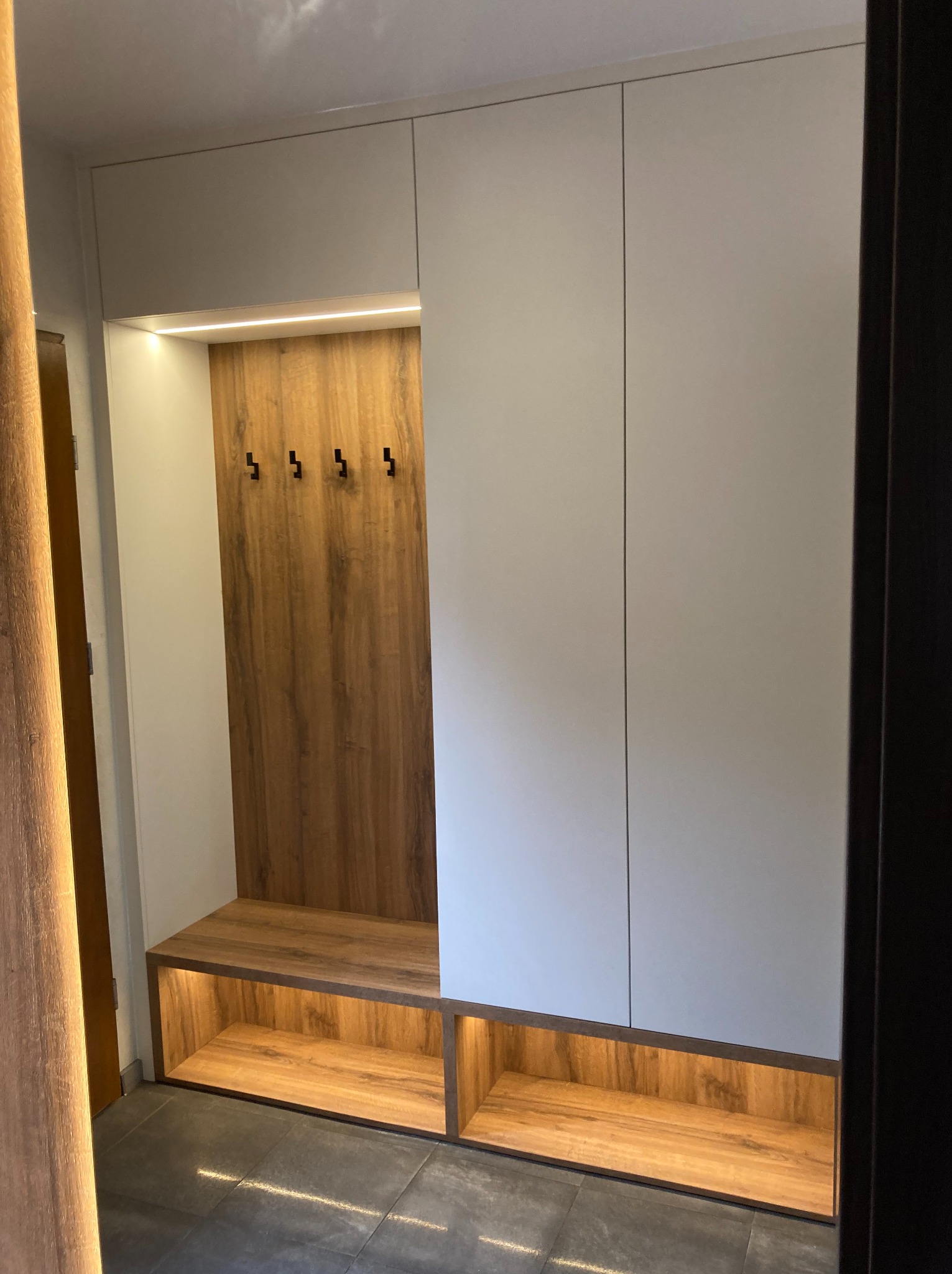Biała szafa wnękowa z drewnianym panelem z haczykami i podświetlaną półką na buty, na tle szarej podłogi, widziana z perspektywy wejścia.