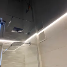 Nowoczesna kabina prysznicowa typu walk-in z deszczownicą, oświetleniem LED wzdłuż ściany i płytkami imitującymi kamień w beżowym odcieniu.