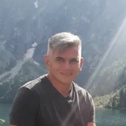 Mężczyzna z siwymi włosami na tle g&oacute;rskiego jeziora i oświetlonych słońcem szczyt&oacute;w.