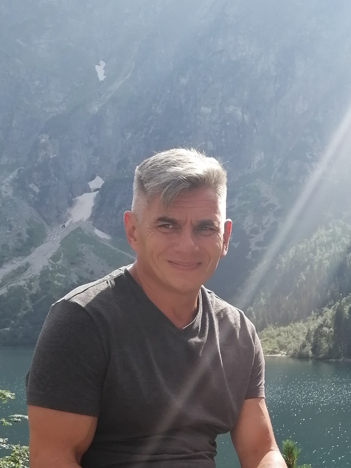 Mężczyzna z siwymi włosami na tle górskiego jeziora i oświetlonych słońcem szczytów.