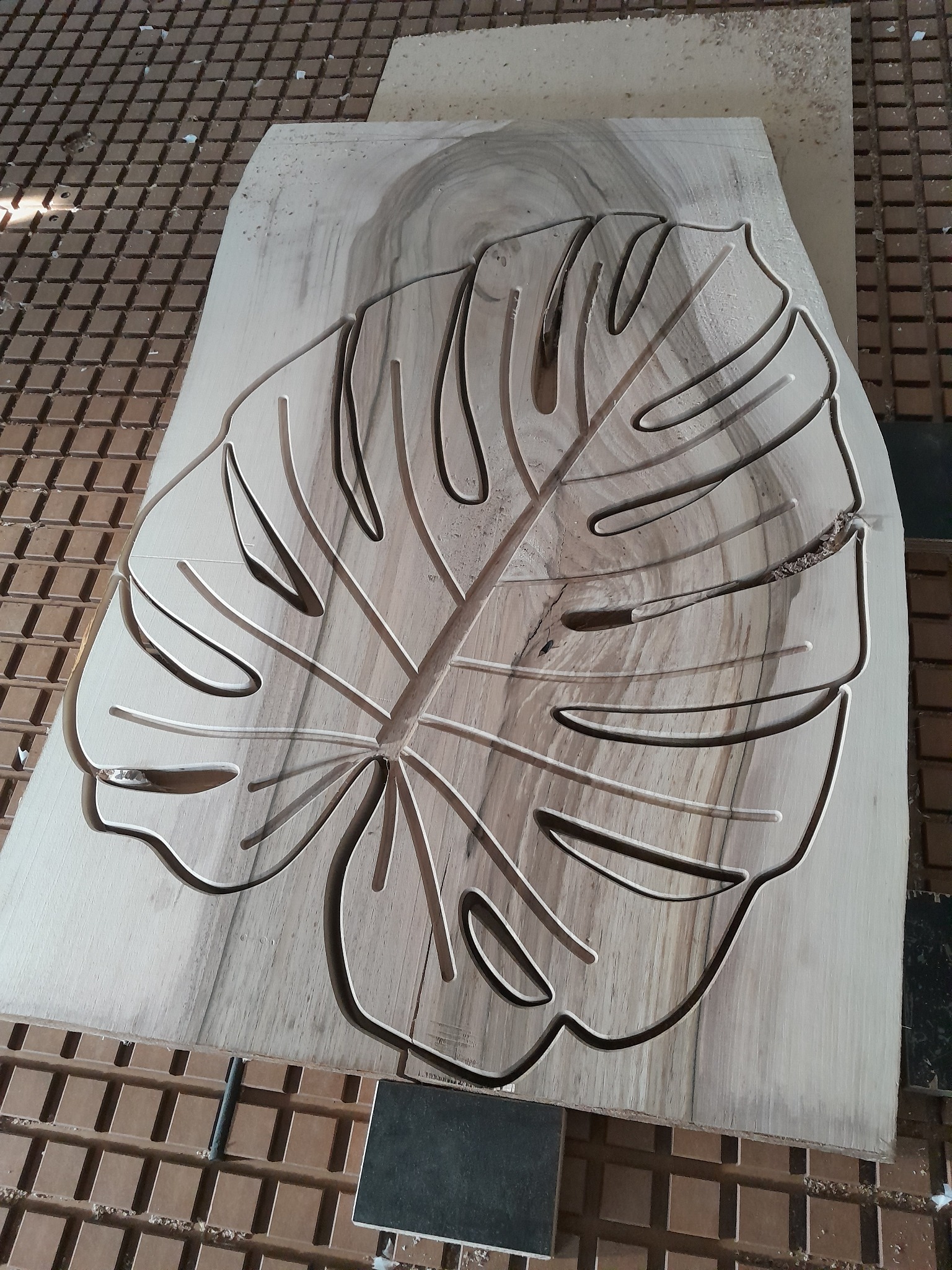 Drewniany panel z wyfrezowanym wzorem liścia monstery, leżący na stole roboczym z widocznymi śladami obróbki.