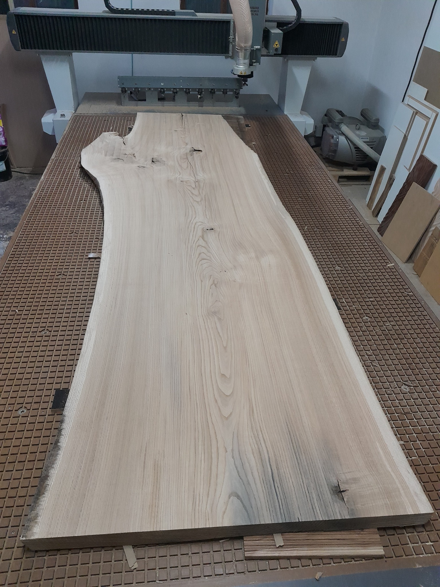 Surowy blat drewniany o nieregularnym kształcie, umieszczony na stole frezarki CNC z widocznymi śladami po frezowaniu.