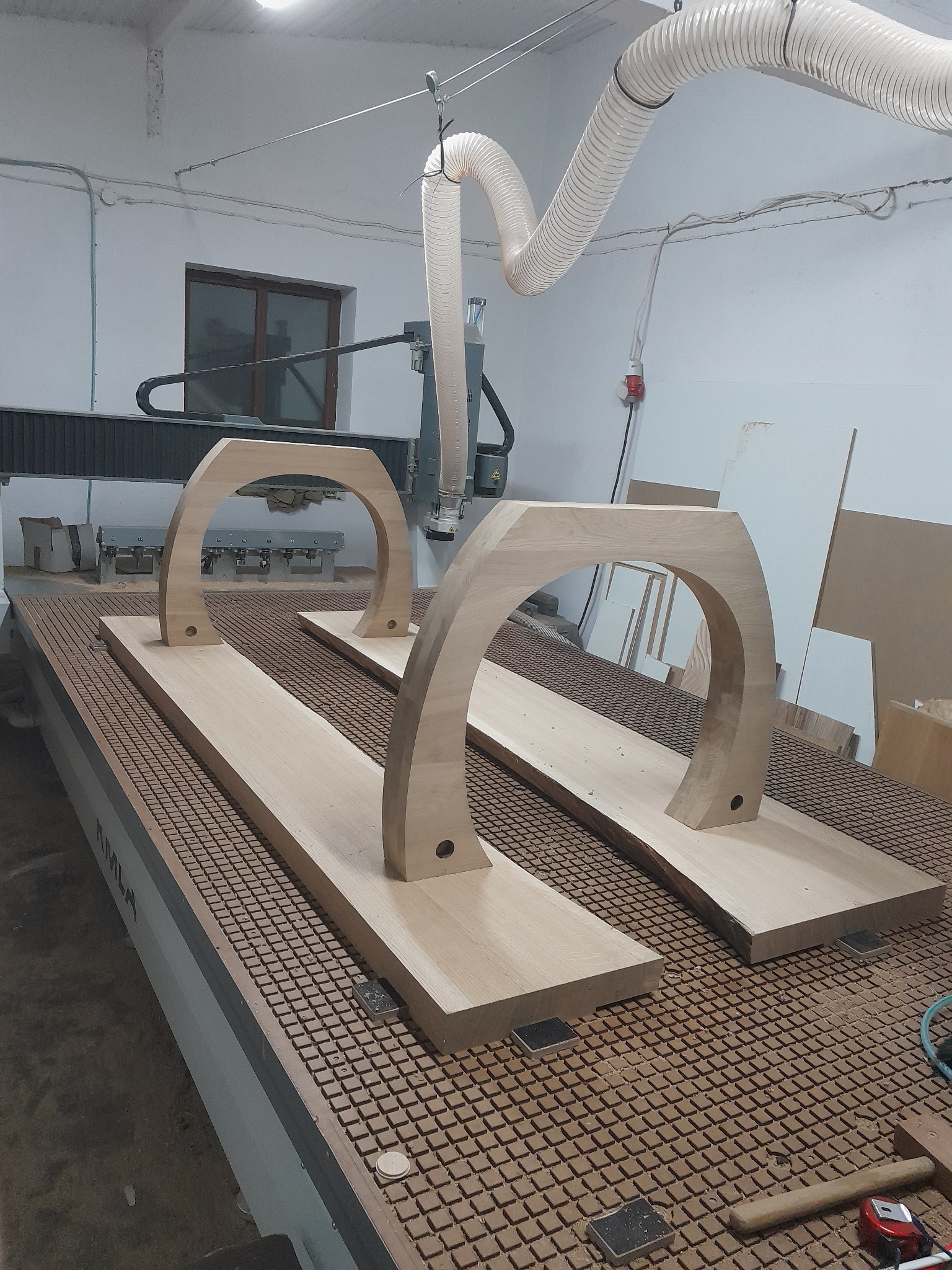 Dwa drewniane elementy w kształcie łuku, z otworami, umieszczone na stole frezarki CNC, z widocznymi śladami obróbki i systemem odsysania pyłu w tle.
