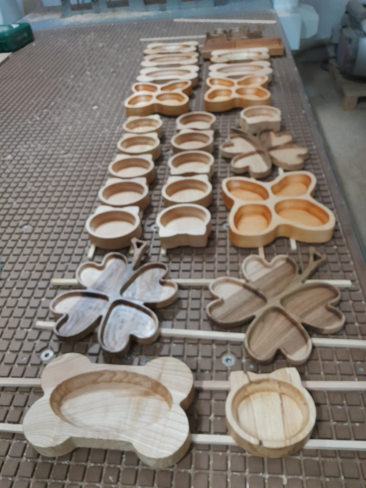 Drewniane tacki i miseczki o różnych kształtach (czterolistna koniczyna, kość, głowa kota) ułożone rzędami na stole z rastrem, po frezowaniu.