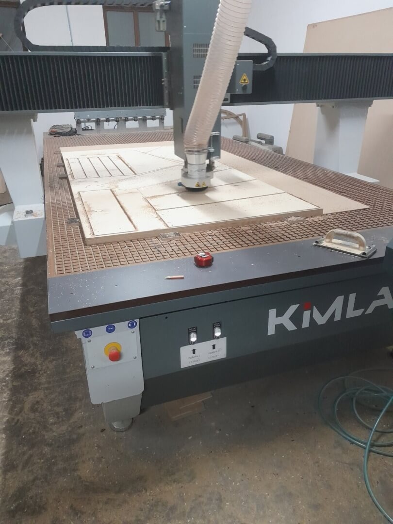 Frezarka KIMLA w trakcie wycinania wzoru z płyty drewnianej, widoczne ślady obróbki i węże odpylające.