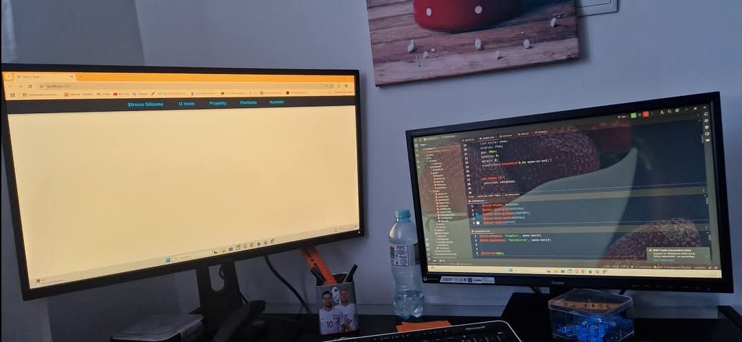 Dwa monitory: na jednym strona www w budowie z menu, na drugim kod źródłowy. Widoczne biurko z akcesoriami i dekoracja ścienna.