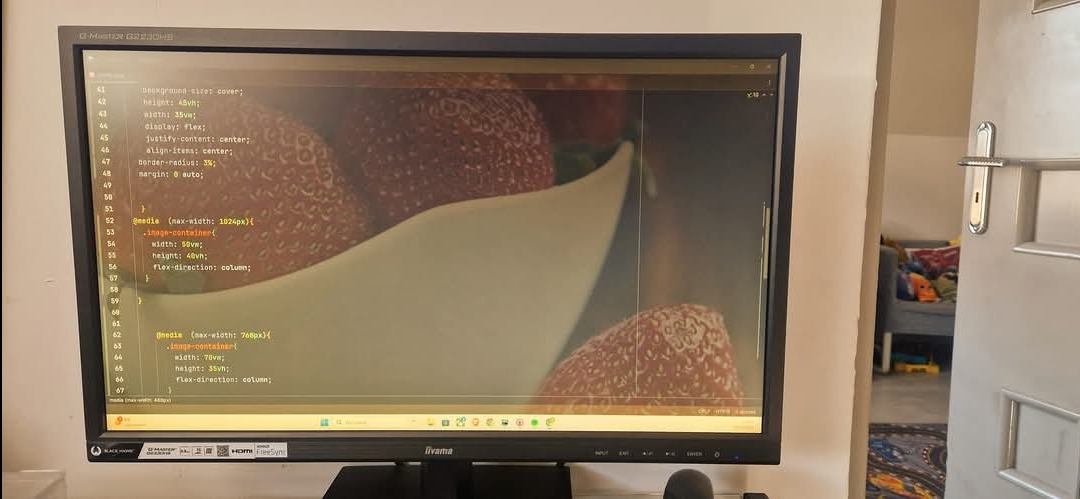 Monitor z kodem CSS i obrazem truskawek. Widoczny fragment pokoju w tle. Prezentacja kodu strony internetowej, być może w trakcie tworzenia lub testowania.