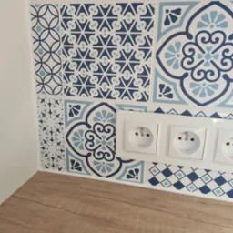 Ściana wyłożona dekoracyjnymi, niebiesko-białymi płytkami ceramicznymi z motywami geometrycznymi i kwiatowymi, z trzema gniazdkami elektrycznymi i fragmentem drewnianego blatu.